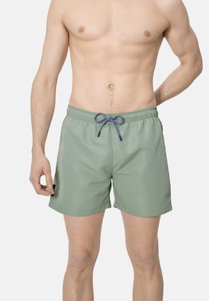 Hombre sin camiseta que lleva pantalones cortos de baño verde claro con cordón azul marino, de pie contra un fondo blanco liso.