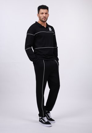 Black athletic tracksuit with white piping details, featuring a zip collar top and matching pants. Model wears black sneakers.