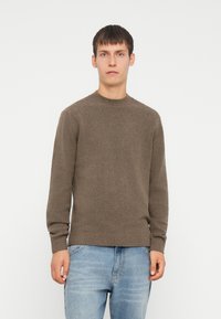 Brązowy sweter dzianinowy z okrągłym dekoltem oraz prążkowanymi mankietami i dołem. Materiał wydaje się miękki i fakturowany, w połączeniu z jasnoniebieskimi dżinsami.