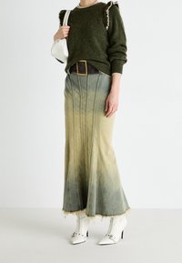 Femme portant un pull vert olive avec des détails à volants, une jupe maxi en denim délavé avec un ourlet effiloché, des bottines blanches à talons et un sac à bandoulière blanc.