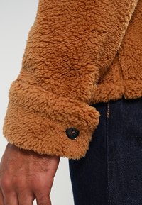 Veste en polaire marron avec un matériau texturé et duveteux, dotée d'un poignet à boutons. La manche s'étend légèrement au-delà du poignet pour davantage de chaleur.