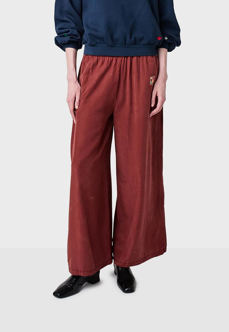 Persoon draagt losse, roestbruine wijde broek met elastische taille en zwarte puntige laarzen, gecombineerd met een marineblauwe blouse met lange mouwen.