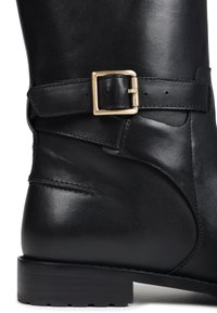 Botte en cuir noir avec une sangle en métal doré près de la cheville et un talon bas empilé sur un fond blanc.