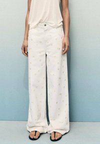 Pantalons larges blancs avec des broderies florales en or, en tissu de coton. Associés à un haut de couleur claire et des tongs noires.