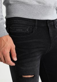 Pantalones vaqueros de mezclilla negra con la rodilla deteriorada y rasgada, un desvanecimiento sutil y una etiqueta con la inscripción "Calvin Klein Jeans". Acompañados de una sudadera gris.