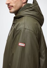 Olīvu zaļa jaku ar kapuci, izgatavota no spīdīgas, ūdensizturīgas materiāla, un ar teksturētu zaļu flīsa apkakli. Piedurknē ir sarkans un balts "HUNTER" logo plāksteris.