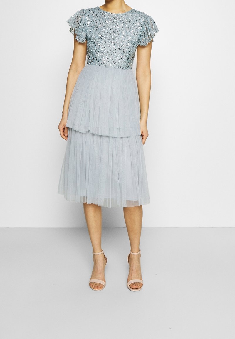 Robe bleu clair avec un corsage pailleté, des manches courtes à volants et une jupe plissée superposée. Portée avec des talons nus à lanières.
