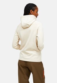 Beige színű kapucnis pulóver puha anyagból, magas gallérral és bordázott mandzsettával, barna nadrággal viselve, látható narancssárga cipzár részletekkel.