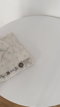 Kokvilnas sega, krēmkrāsā ar gaiši zilu kvadrātu rakstu. Rūpīgi salocīta ar redzamu aprūpes etiķeti, novietota uz apļveida balta galda.