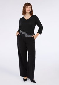 Schwarzer V-Ausschnitt-Pullover mit Dreiviertelärmeln, kombiniert mit schwarzen Hochbundhosen, die einen grau gestreiften Gürtel haben. Schwarze spitze Pumps.