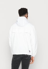 Biała lekka kurtka z kapturem, wyposażona w elastyczne mankiety i sznurek ściągający u dołu. Z logo na dolnej stronie oraz wentylowanymi plecami.