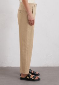 Pantaloni beige a sigaretta in cotone con tasche laterali e pieghe frontali, abbinati a sandali neri con dettaglio in oro sulla fibbia.