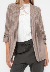 Brązowa marynarka w kratę o luźnym kroju, z wyciętymi klapami i podwiniętymi rękawami, zestawiona z białym, teksturowanym topem i szarymi spodniami.