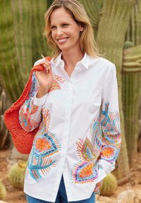 Chemise blanche à boutons avec des motifs floraux colorés en bleu, orange et vert sur les manches. Accompagnée d'un sac orange texturé.