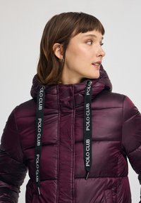 Lila puffjacka med huva, med svarta dragskor märkta "POLO CLUB." Tillverkad av glänsande nylon med ett vadderat mönster.
