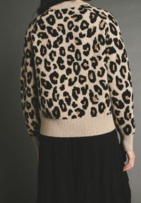 Personne portant un pull beige à imprimé léopard avec des poignets et un ourlet côtelés, associé à une jupe noire texturée, vue de dos.