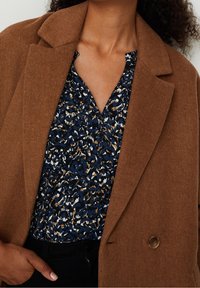 Brązowa marynarka z mieszanki wełny o teksturowanym wykończeniu, zestawiona z granatową wzorzystą bluzką z abstrakcyjnymi kształtami w kolorze kremowym i jasnobrązowym.