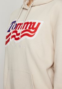 Sweat à capuche beige avec un patch logo "Tommy Jeans" rouge, blanc et bleu et une poche avant, porté par une personne de l'épaule à la taille.