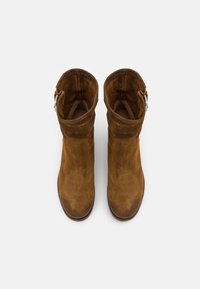 Botines de ante marrón con un eje ligeramente ajustado, punta redondeada y un acento de hebilla plateada a un lado. Textura suave y acabado mate.