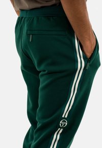 Pantalones de chándal verdes hechos de tejido suave, con rayas laterales blancas, un bolsillo trasero y un parche de logo cerca del dobladillo.