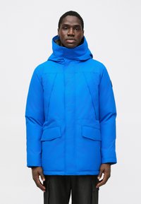 Blue insulated jacket with a high collar and hood. Features large front pockets and a smooth, shiny finish. Overall, a structured design.