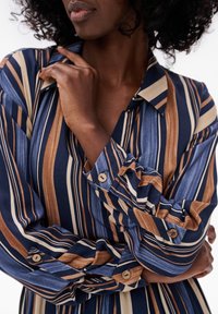 Bluzka w paski w kolorach niebieskim, beżowym i brązowym. Długie rękawy z zapięciami na guziki, kołnierzyk oraz gładka tekstura materiału.
