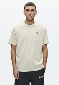 Beige bomulds t-shirt med rund hals og korte ærmer. Har et lille blåt logo på den øvre venstre bryst. Båret med sorte bukser.