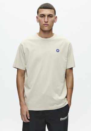 Beige katoenen t-shirt met een ronde hals en korte mouwen. Heeft een klein blauw logo op de bovenste linkerkant van de borst. Draagt met zwarte broek.