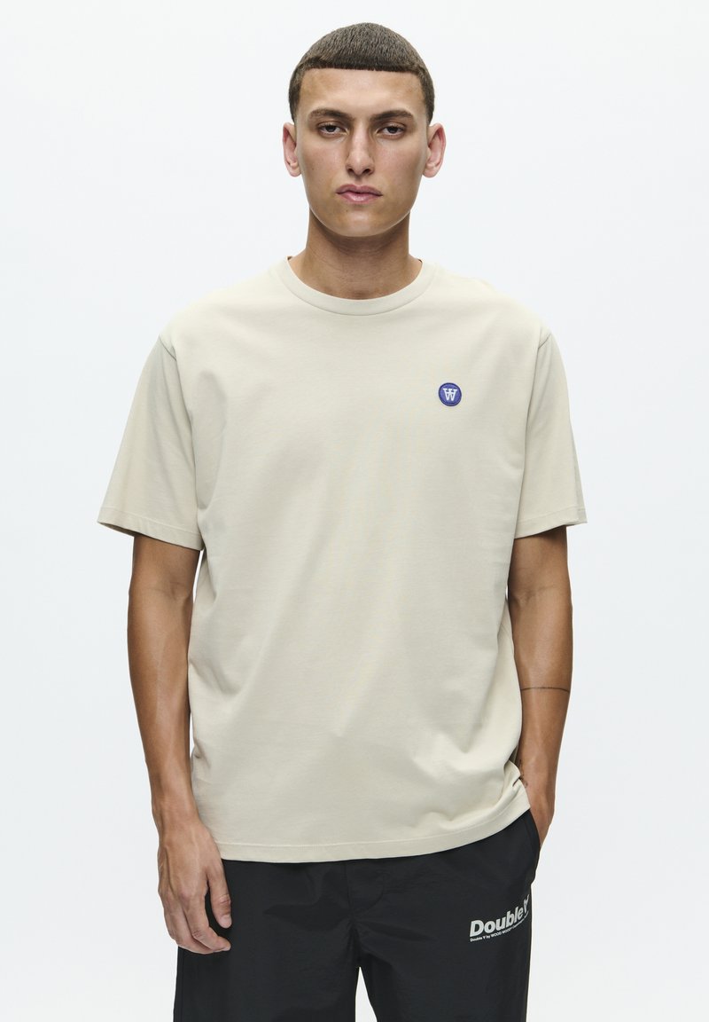 Beige bomulds t-shirt med rund hals og korte ærmer. Har et lille blåt logo på den øvre venstre bryst. Båret med sorte bukser.