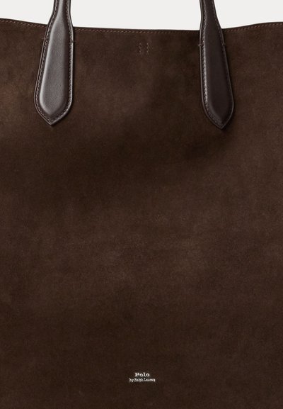 Sac fourre-tout en suède brun avec des poignées en cuir foncé, présente un imprimé logo en bas, un design simple et une texture douce.