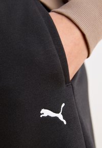 Čierna tkanina s hladkou textúrou, s bielym logom Puma. Z čiernej kapsy vyčnieva béžový rukáv.