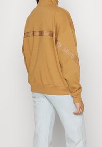 Sweatshirt brun à col montant, avec une bande logo ton sur ton dans le dos. Texture douce, coupe décontractée et manches longues.