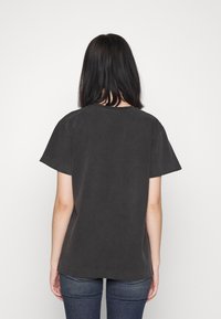 Osoba z włosami do ramion w ciemnym kolorze, ubrana w prostą czarną koszulkę z krótkim rękawem i ciemnoniebieskie dżinsy, stająca tyłem na białym tle.