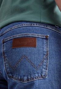 Jeans de denim azul con un parche de cuero marrón de la marca que presenta el logo "Wrangler", destacando un diseño de costuras distintivas y bolsillos.