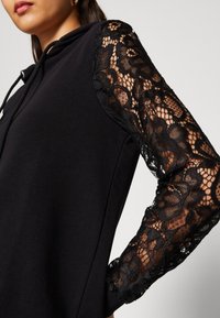 Top nero con cappuccio, caratterizzato da maniche lunghe in pizzo nero intricati, con motivi floreali lungo il braccio e un corpo in tessuto liscio.