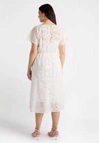 Weißes, knielanges Kleid mit floralem Lochmuster, kurzen Ärmeln und elastischem Bund. Getragen mit beigen Riemchensandalen.