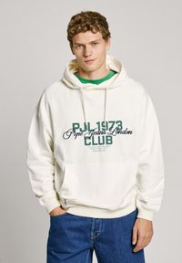 Felpa crema con stampa frontale "PJL 1973 Pepe Jeans London CLUB" in verde e blu navy. Presenta una tasca a marsupio e polsini e orlo a costine. Jeans blu.