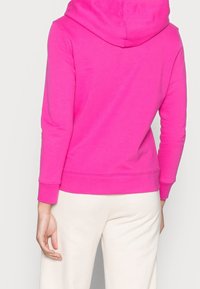 Felpa rosa con cappuccio regolabile, maniche lunghe e polsini a costine. Texture morbida, design minimalista, vista dal retro, abbinata a pantaloni chiari.