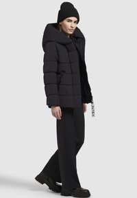 Schwarze gepolsterte Jacke mit großer Kapuze, horizontalem Steppmuster, seitlichen Reißverschlüssen und einem Etikett-Detail. Getragen mit schwarzen weit geschnittenen Hosen und Stiefeln.