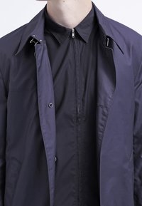 Veste en tissu bleu marine au design épuré, dotée d'une fermeture à boutons, d'un devant zippé, d'un col pointu et d'une texture élégante.