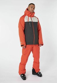 Orange Skijacke mit grauen und weißen Akzenten, ausgestattet mit einem Reißverschluss vorne und einer Kapuze. Kombiniert mit passenden orangen Skihosen und schwarzen Skischuhen.