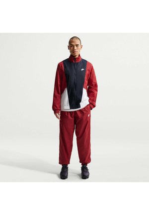Mann steht in einem roten, schwarzen und weißen Nike-Trainingsanzug mit schwarzen Sneakers auf einem schlichten hellen Hintergrund.