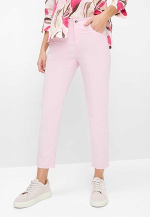 Frau trägt hellrosa eng anliegende Jeans, weiße Turnschuhe und ein Hemd mit Blumenmuster, steht mit einer Hand in der Tasche.