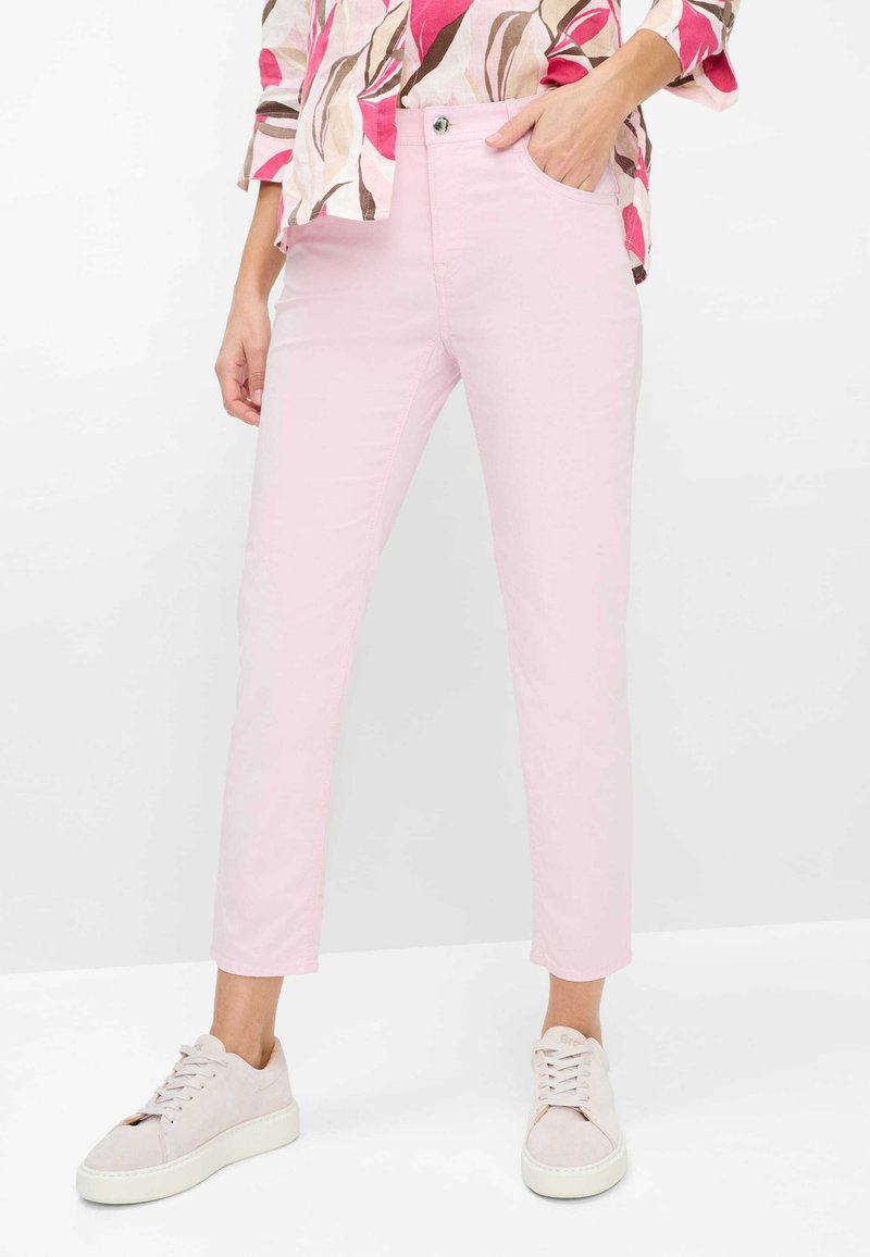 Kvinde iført lyserøde, tætsiddende jeans, hvide sneakers og en blomstret skjorte, stående med den ene hånd i lommen.