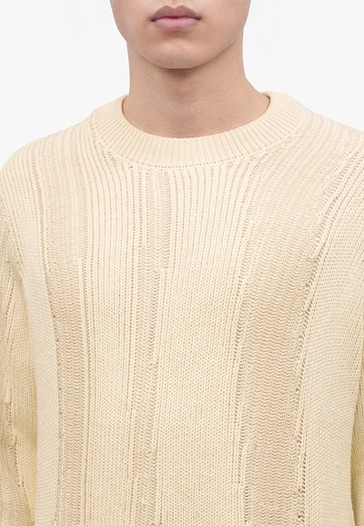 Gros plan d'une personne portant un pull tricoté couleur crème avec une texture côtelée et un col rond.