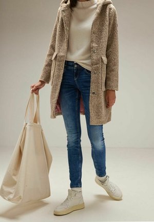 Osoba ubrana w beżowy płaszcz typu teddy, biały sweter, niebieskie dżinsy, białe trampki, trzymająca dużą beżową torbę typu tote w neutralnym otoczeniu.