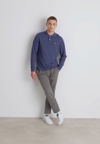 Marineblaues Langarm-Polohemd mit Kragen, graue schmal geschnittene Hose und weiße Sneaker, gelehnt gegen eine helle Wand auf einem neutralen Boden.