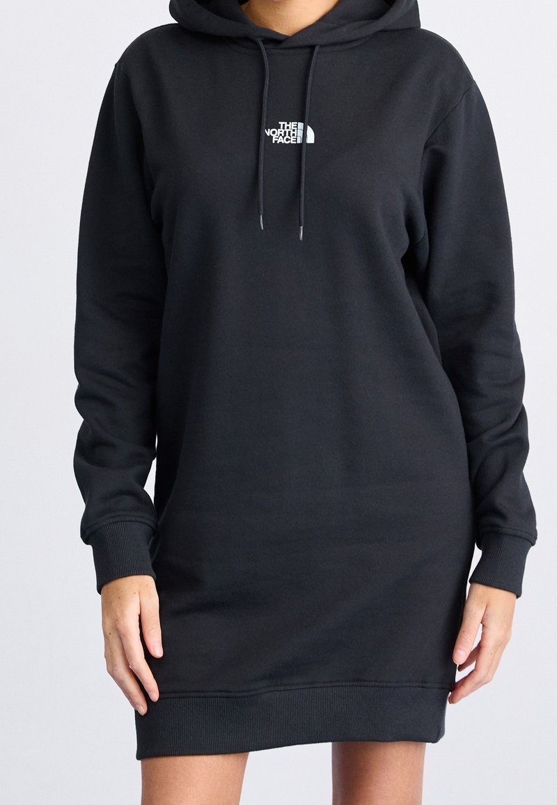 Personne portant une robe-hoodie longue noire avec un logo blanc "The North Face" à l'avant et des cordons sur la capuche, les mains posées naturellement le long du corps.