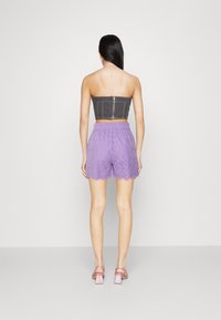 Femme debout, de dos, vêtue d'un crop top gris sans bretelles, d'un short en broderie anglaise lavande et de sandales à talons blocs irisés sur fond uni.