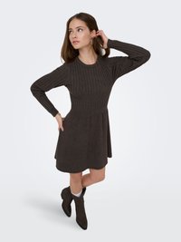 Robe en maille côtelée brun foncé avec manches longues, corsage ajusté et jupe évasée. Assortie à des bottines foncées et des boucles d'oreilles créoles.